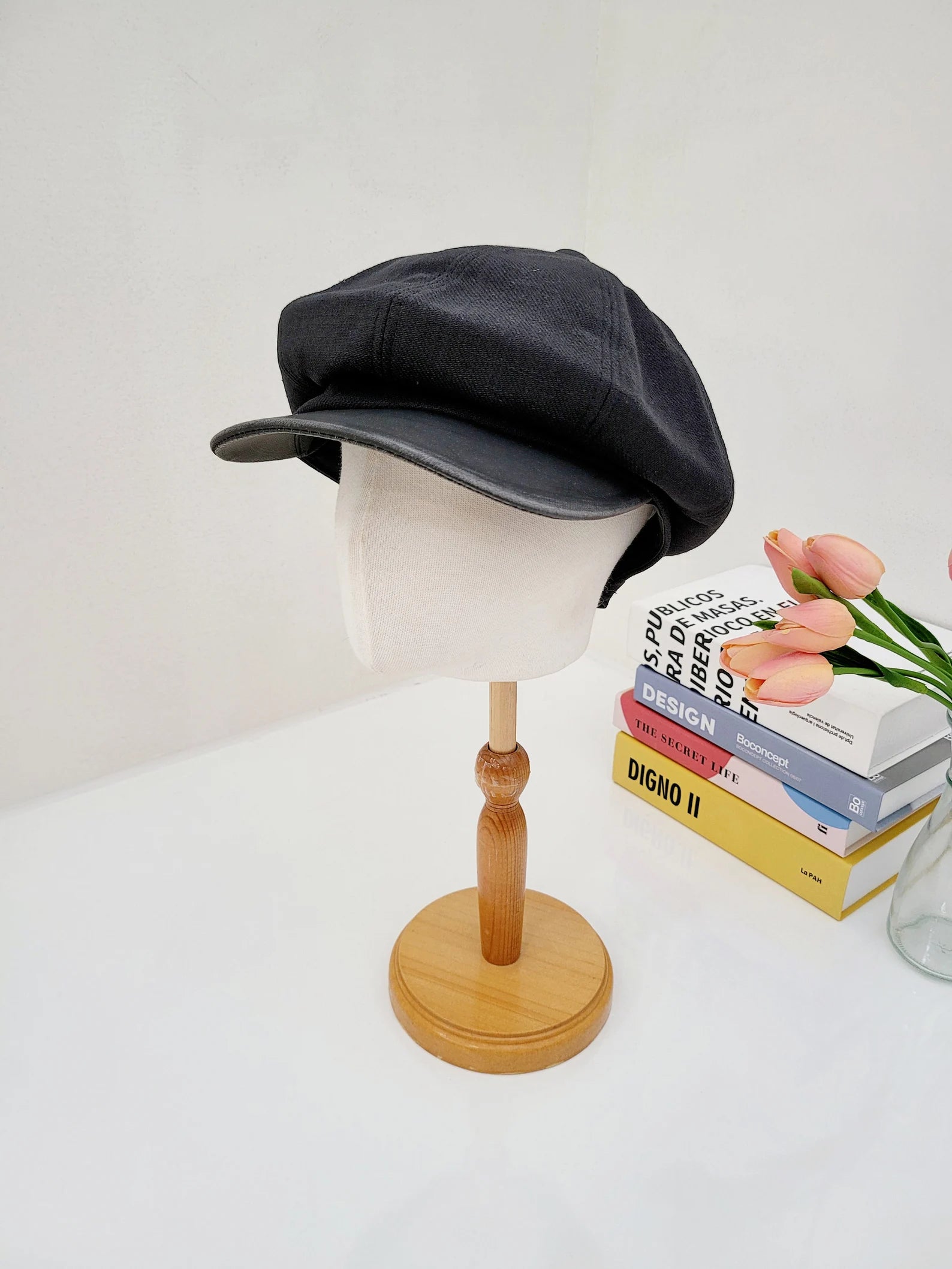 Black lana beret on a mannequin head, atop a wooden stand. Baker boy style hat is displayed next to books and pink tulips.