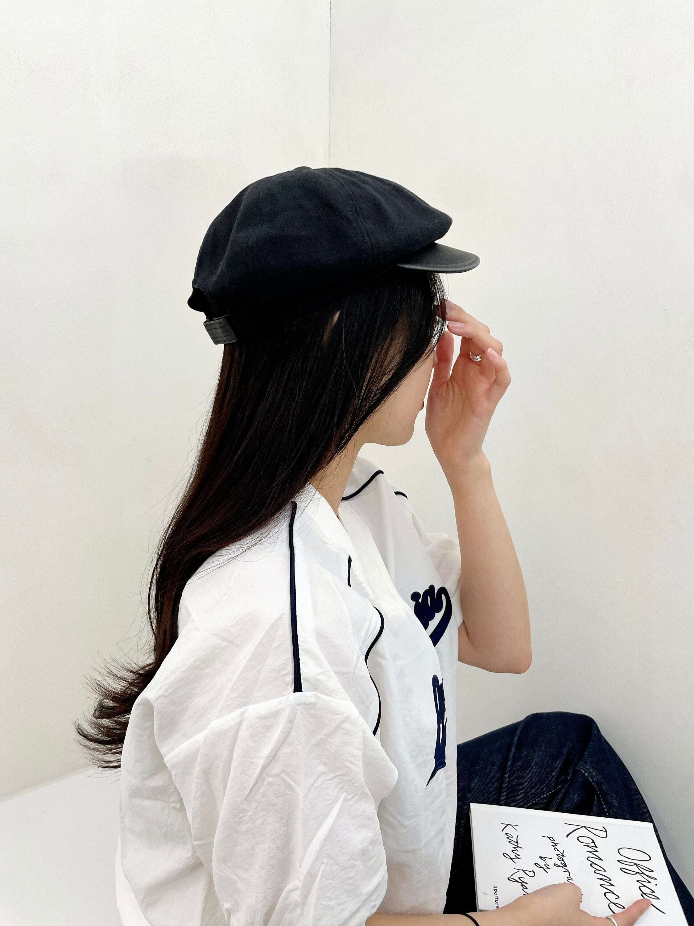 Woman in profile wearing a black lana beret and white shirt, holding a book. The beret has a classic, slightly puffed design.
