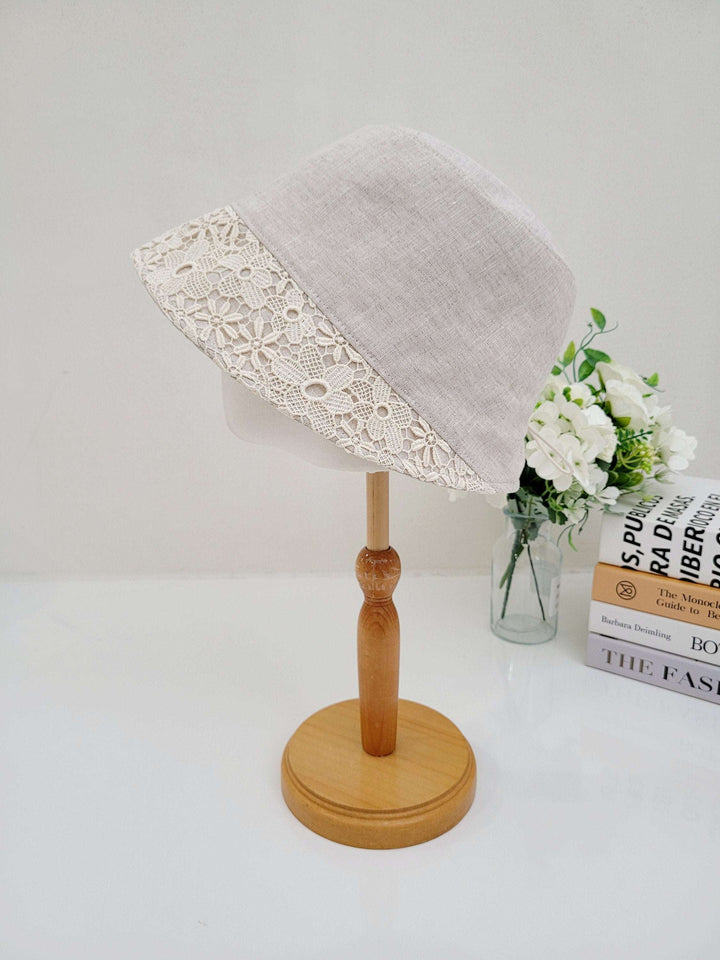 Linen bucket-flower eyelet hat in neutral linen with a delicate floral lace trim, displayed on a wooden stand.