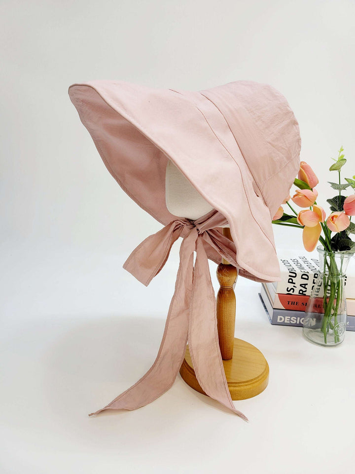 my fair lady: Soft pink sun hat with a wide, floppy brim and long ties, displayed on a stand with flowers in the background.