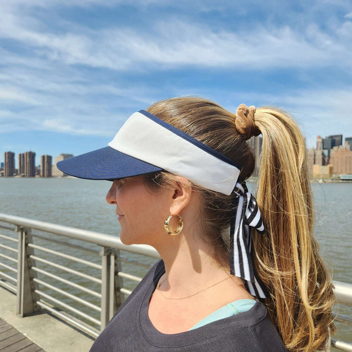 Navy and white lic visor with a striped ribbon detail, worn with a blonde ponytail. Cityscape backdrop.