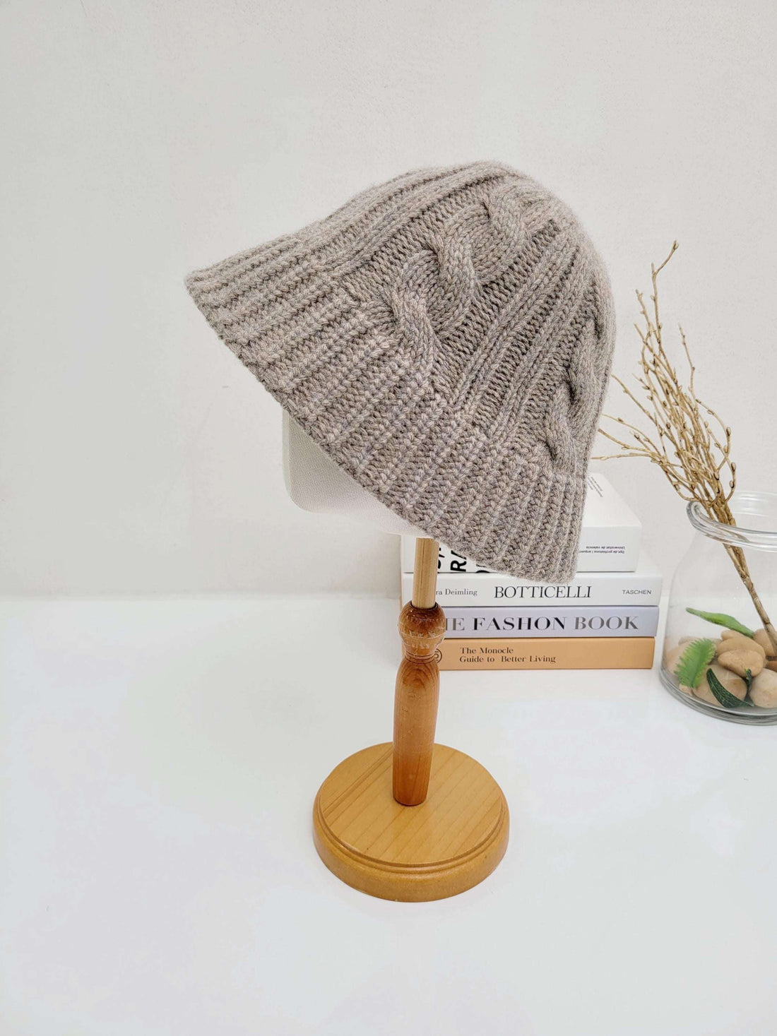 Beige cable knit hat displayed on a wooden stand, showcasing its intricate knit pattern and soft texture. A stylish and cozy accessory.