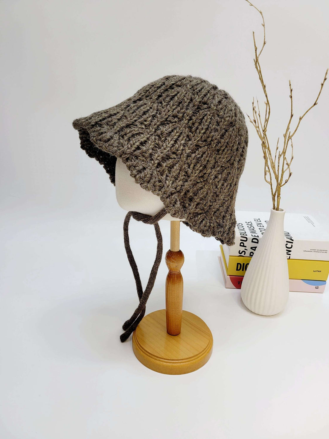 Braided wool hat in earthy brown, featuring a textured knit and tie closures, displayed on a wooden stand