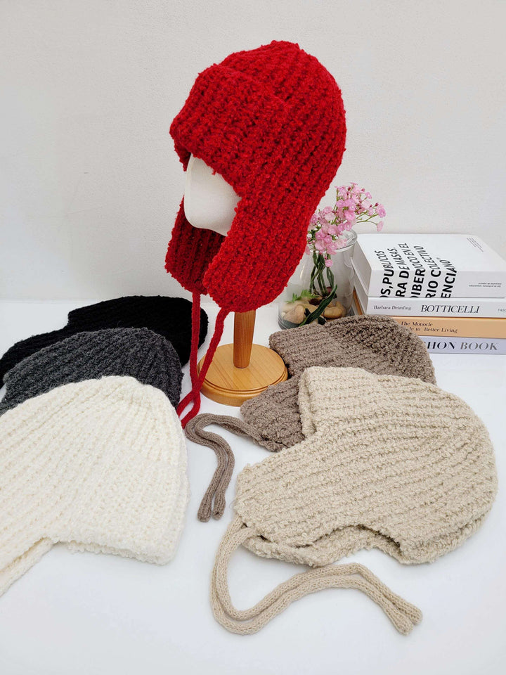 Red knitted wool earflap hat on a mannequin, with similar hats in white, gray, brown, and black around it. Warm and stylish winter headwear.