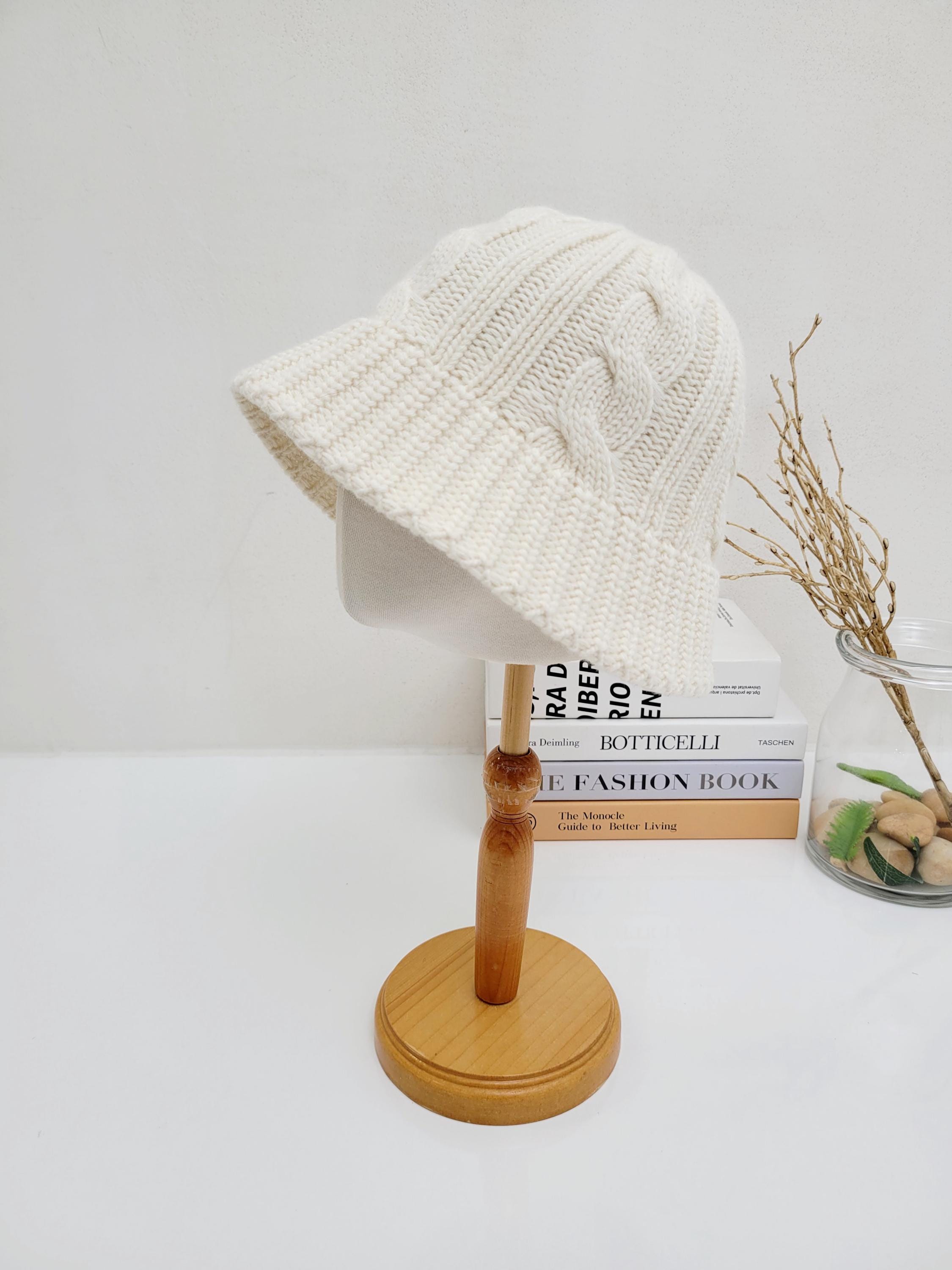 Cream cable knit hat on a wooden stand. Cozy knit bucket hat with a classic cable pattern.