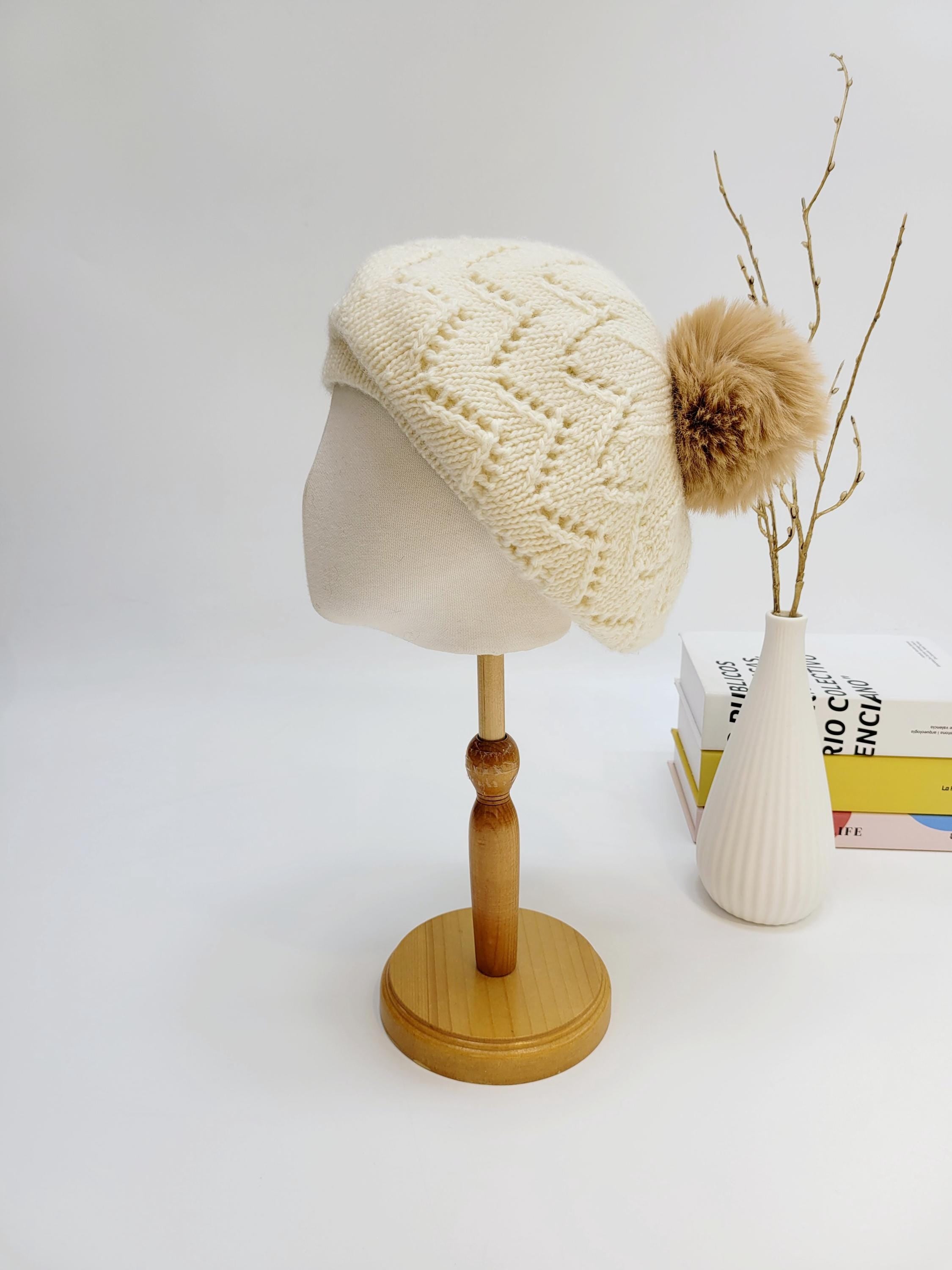 Cream knitted beret, an emma in paris design, with geometric pattern and brown pompom, displayed on a wooden stand.