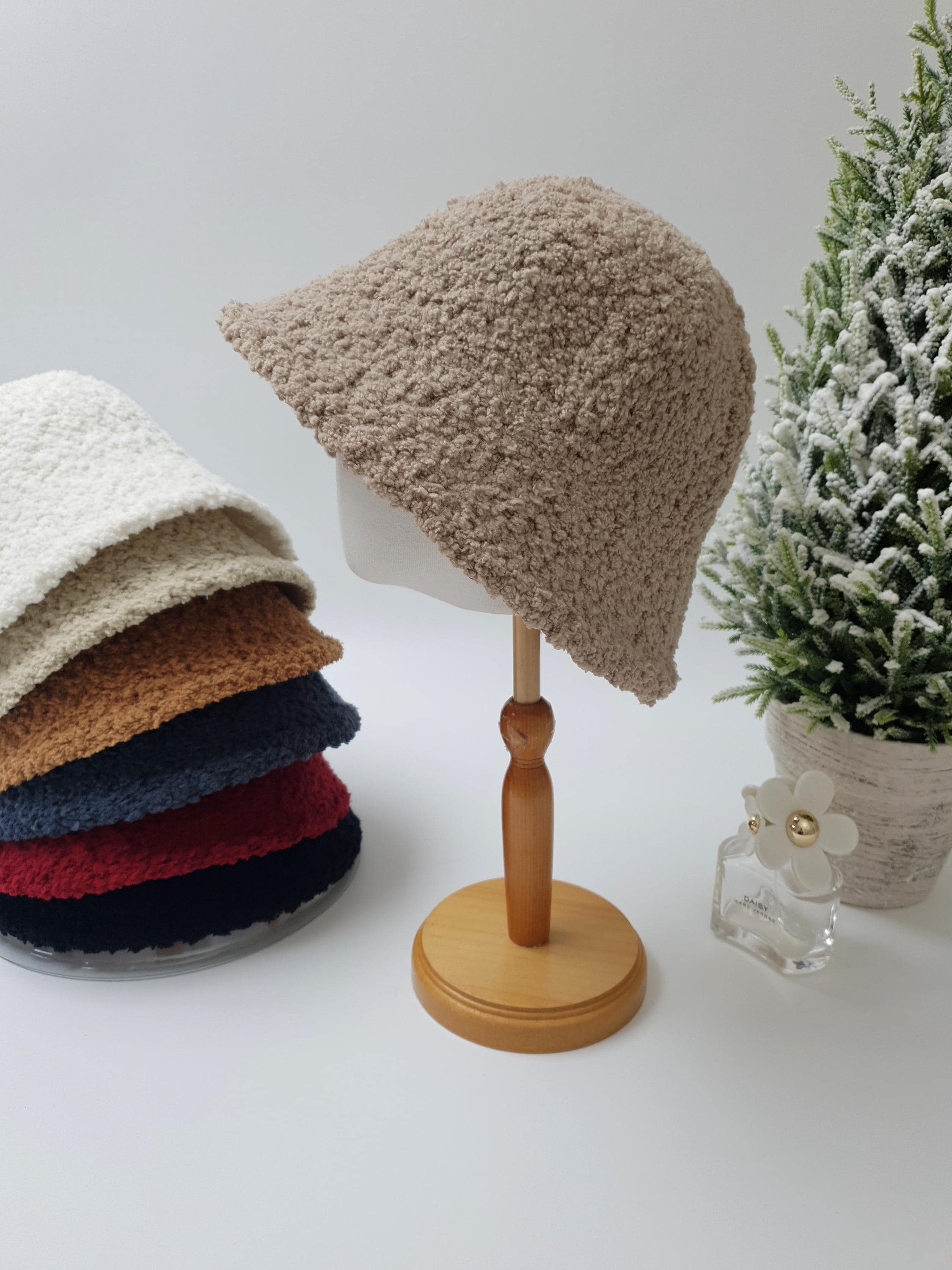 Beige sherpa bucket hat on a wooden stand. Stack of sherpa bucket hats in white, brown, navy, red, and black visible. Festive decor includes mini Christmas tree.