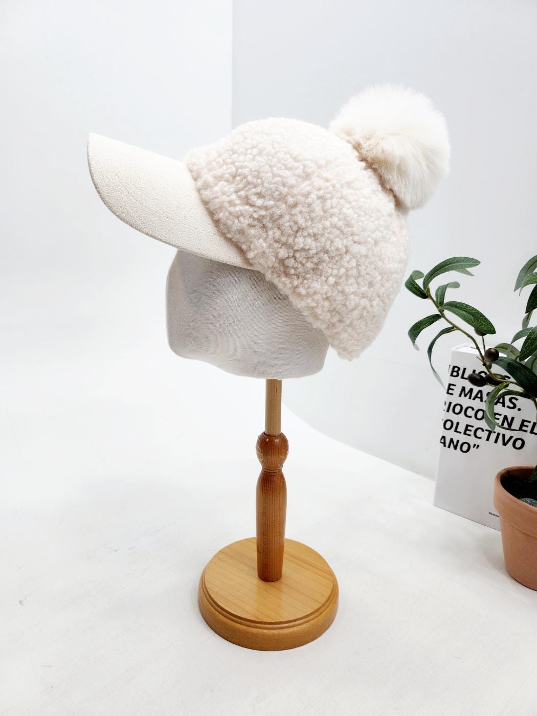Creamy white adriana sherpa cap with a solid bill and fluffy pom-pom, displayed on a wooden stand.