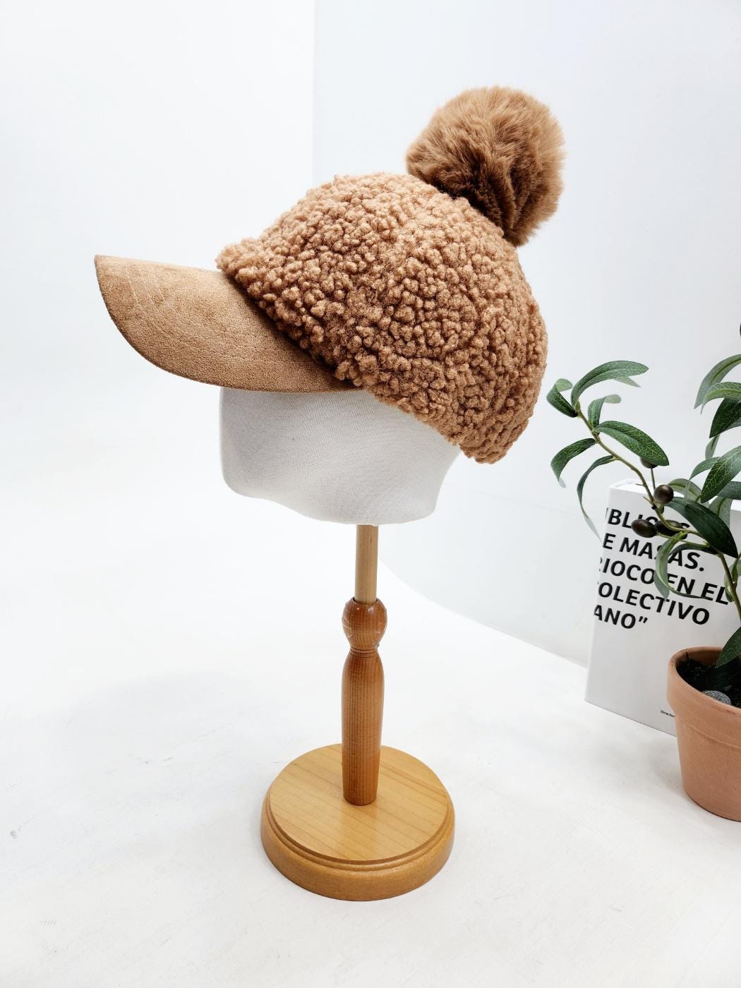 ADRIANA SHERPA CAP: Brown sherpa hat with a suede brim and a fluffy pom-pom on a stand