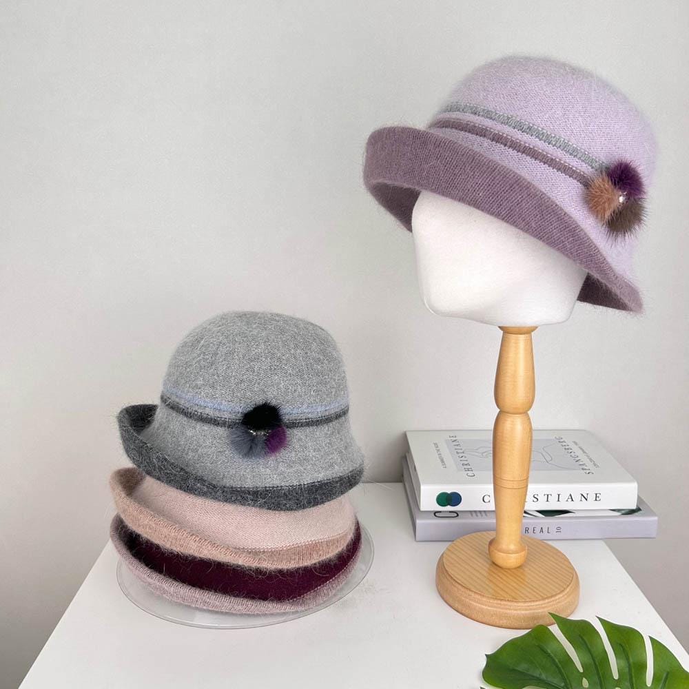 Angora elizabeth hat: lavender cloche on a stand, plus a stack of gray, pink, and burgundy hats, each with a dark band and pompom detail.